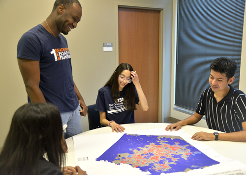 Four students collaborating around a large map on a table in a classroom. One student stands, smiling and looking at the map, while the others are seated, examining the details of the map. They appear to be engaged in a discussion about the map's data. The students are wearing UTSA t-shirts, indicating their affiliation with the Urban and Regional Planning program.