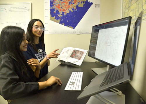 Two students working together at a desk with dual computer monitors displaying a detailed map. One student is pointing at the screen while holding an open textbook, which also contains map-related information. Both students are focused on analyzing the map data, surrounded by wall posters displaying additional maps and diagrams, indicating a collaborative learning environment in the Urban and Regional Planning program at UTSA.