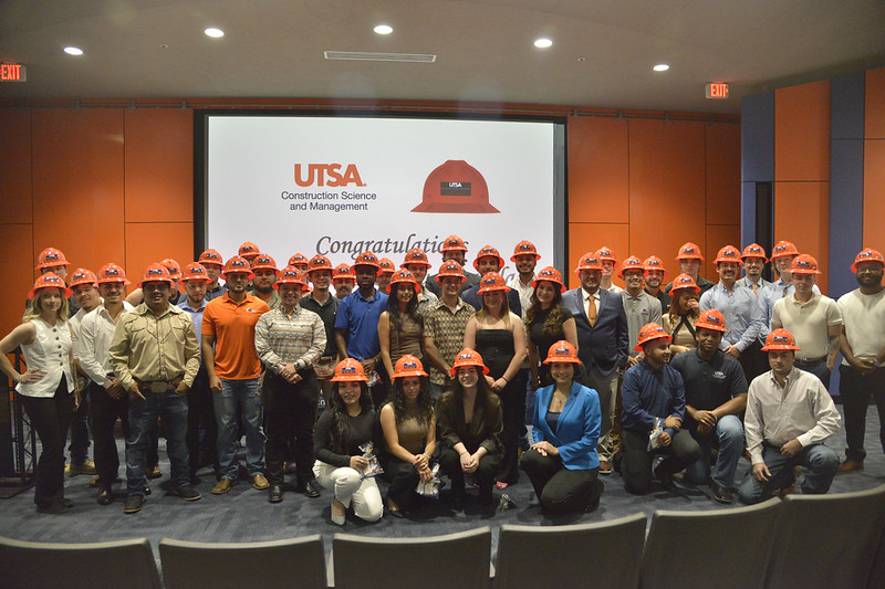 A large group of Construction Science and Management students and faculty from UTSA's Klesse College pose together in an auditorium wearing bright orange hard hats during the Spring 2025 Hard Hat Ceremony.