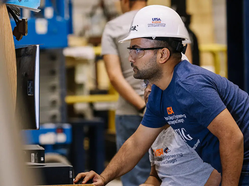 Student in a UTSA hard hat works at a computer in an industrial lab, highlighting problem-solving and efficiency in Industrial and Systems Engineering.