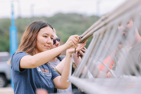 A student wearing a UTSA Klesse College t-shirt works with a team to assemble a steel bridge structure outdoors.