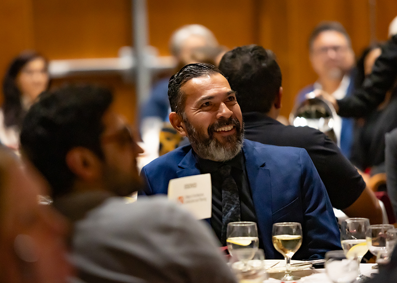 Armando smiles and looks engaged while seated at a banquet table during the AIA San Antonio awards event. Dressed in a blue suit, he sits among other guests, with glasses of white wine and a warm event ambiance in the background.