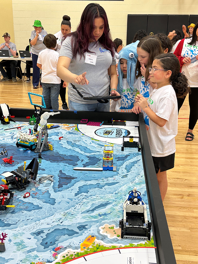Indra Garcia-Morales kneels beside a table, demonstrating a robotics activity to a group of engaged elementary students during the NISD LEGO League Challenge.
