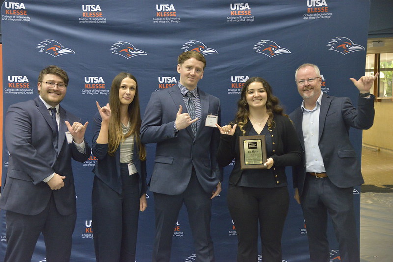 Five people in business attire stand in front of a UTSA Klesse College of Engineering and Integrated Design backdrop, smiling and making the "Birds Up" hand sign. One person holds a plaque reading "1st Place."