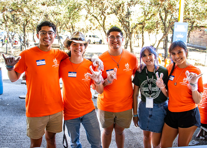 Five UTSA students smile and pose with "Birds Up" hand signs at an outdoor volunteer event