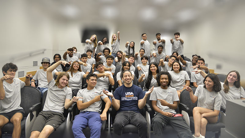 Participants of the Klesse College Summer Bridge Program gathered in an auditorium with a faculty member at the center, all smiling and pointing, showcasing the sense of community and connections built during the program. The diverse group reflects the supportive environment that continues throughout their freshman year.