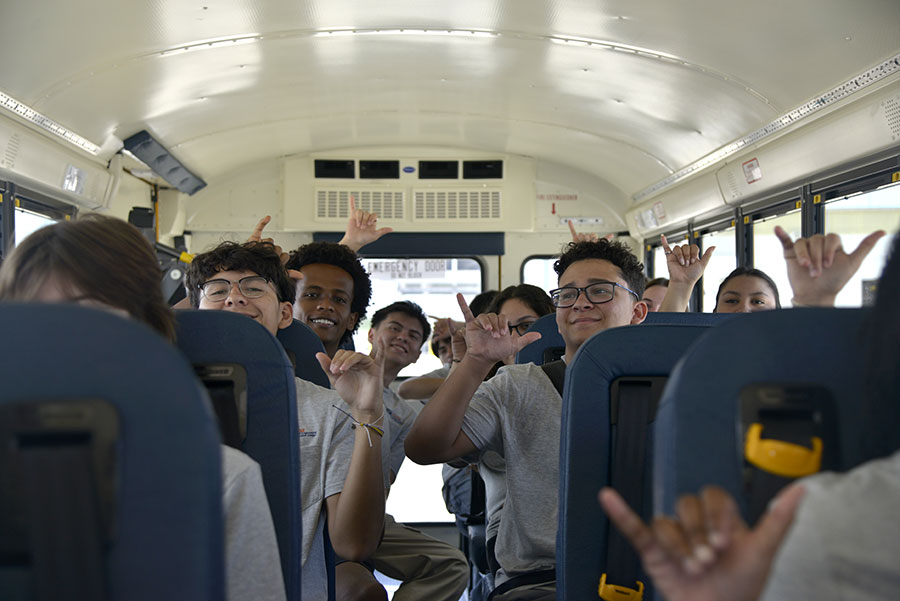 Students on a school bus smiling and making hand gestures, symbolizing camaraderie and excitement during the Klesse College Summer Bridge Program.