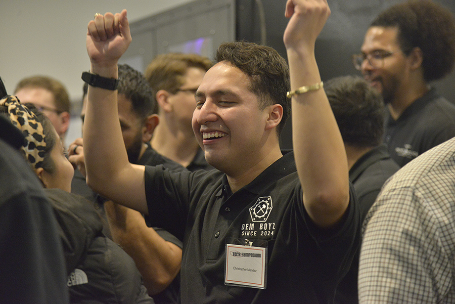 A smiling member of the third-place team, DEM Boys Engineering Group, celebrates at the Fall 2024 Tech Symposium hosted by the Margie and Bill Klesse College of Engineering and Integrated Design. The individual, wearing a black polo shirt with the team name 'DEM Boys Since 2024,' raises both arms in victory amid a crowd of other participants and attendees.